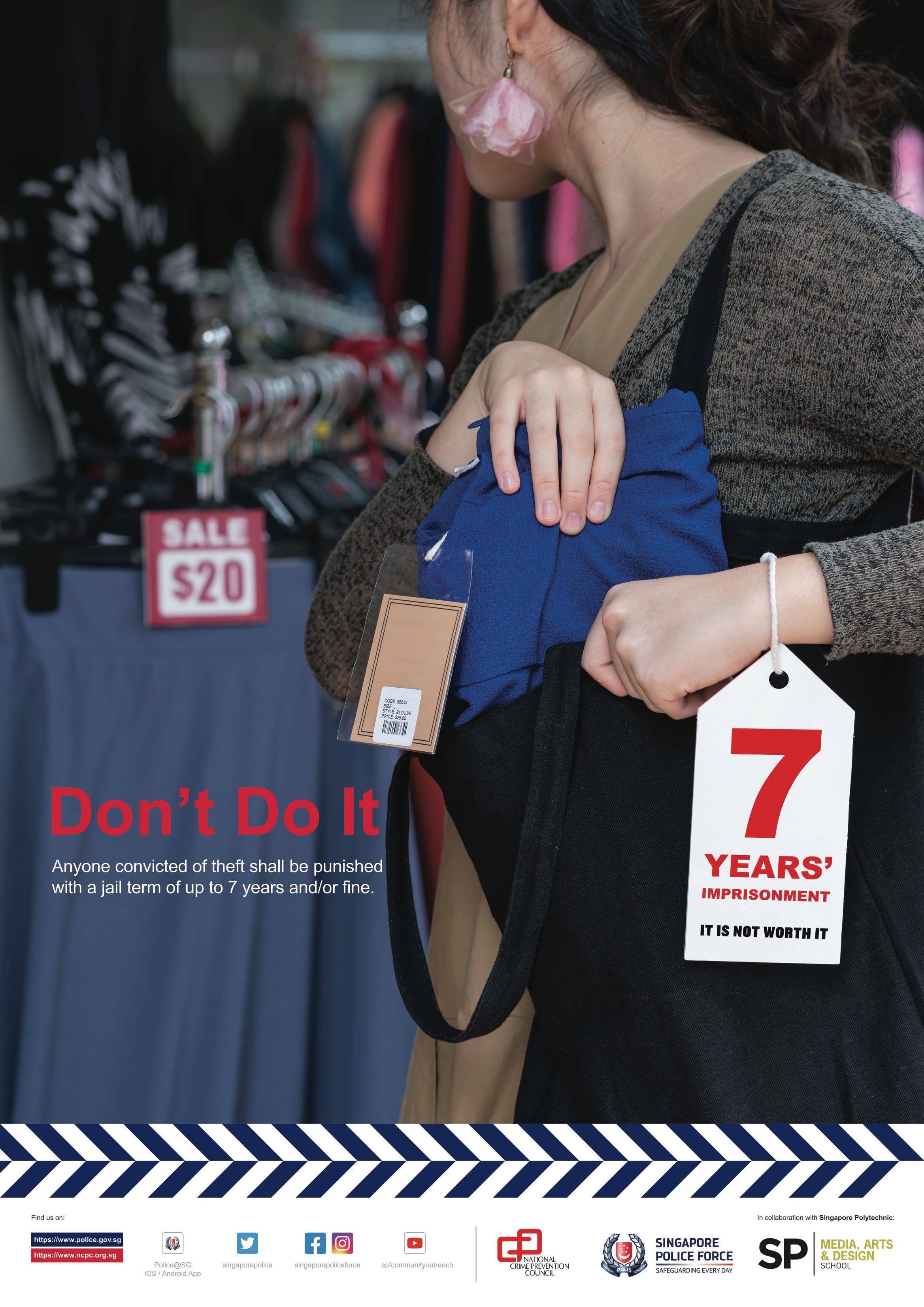 A close-up framing shows a person in a general retail store setting placing a blue garment or fabric item with a price tag into a black tote bag, with a hanging advisory tag featuring red typography that reads "7 YEARS' IMPRISONMENT IT IS NOT WORTH IT".